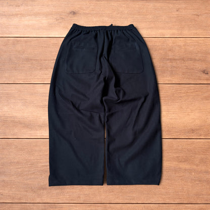 Black baggy wool trousers laid flat on wooden floor showing elasticated waistband and pockets.