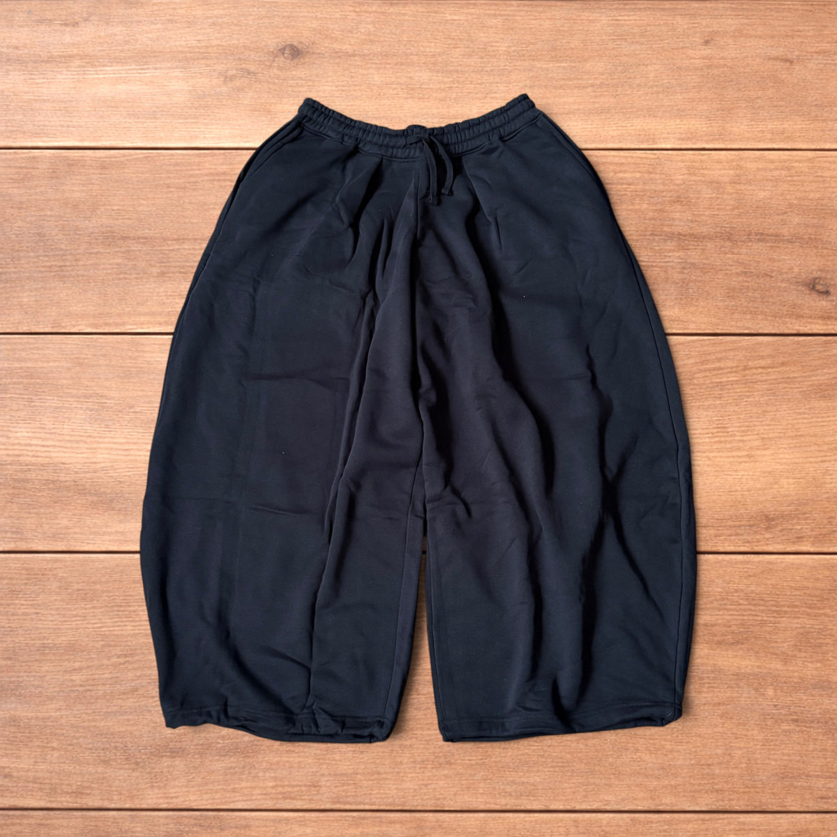 Black baggy joggers laid flat on wooden surface showing elasticated waistband, drawstring and elasticated ankle hems.