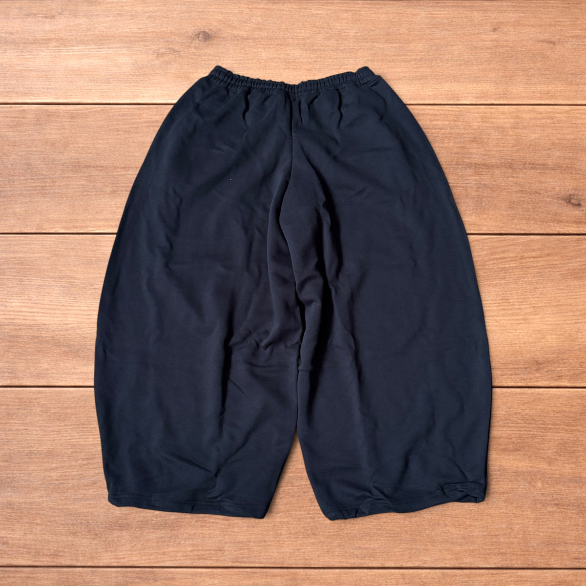 Black baggy joggers laid flat on wooden surface showing elasticated waistband, drawstring and elasticated ankle hems.