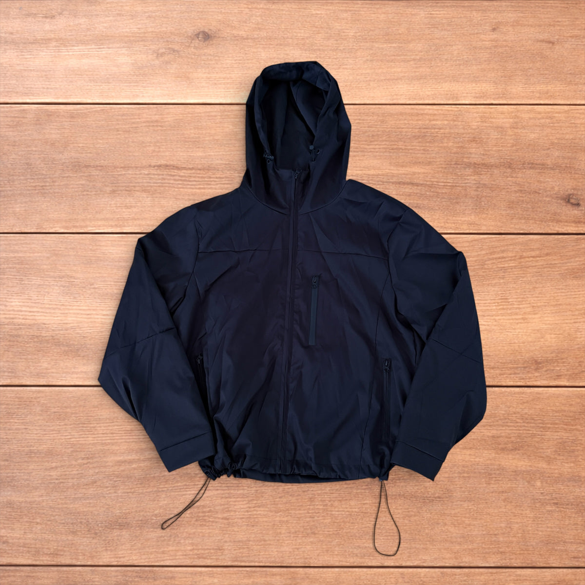 Black outdoor windbreaker jacket with hood, zip pockets and toggled hem laid flat on wooden surface.