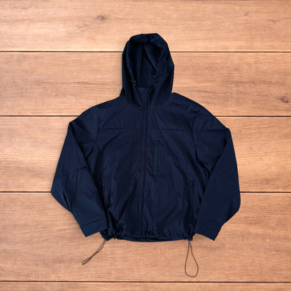 Black outdoor windbreaker jacket with hood, zip pockets and toggled hem laid flat on wooden surface.