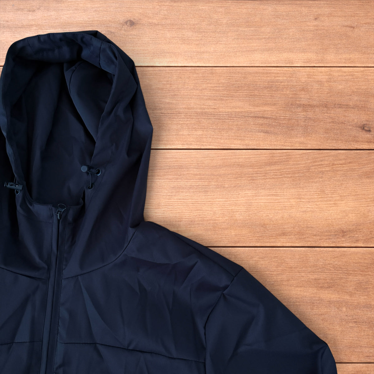 Black outdoor windbreaker jacket with hood, zip pockets and toggled hem laid flat on wooden surface.