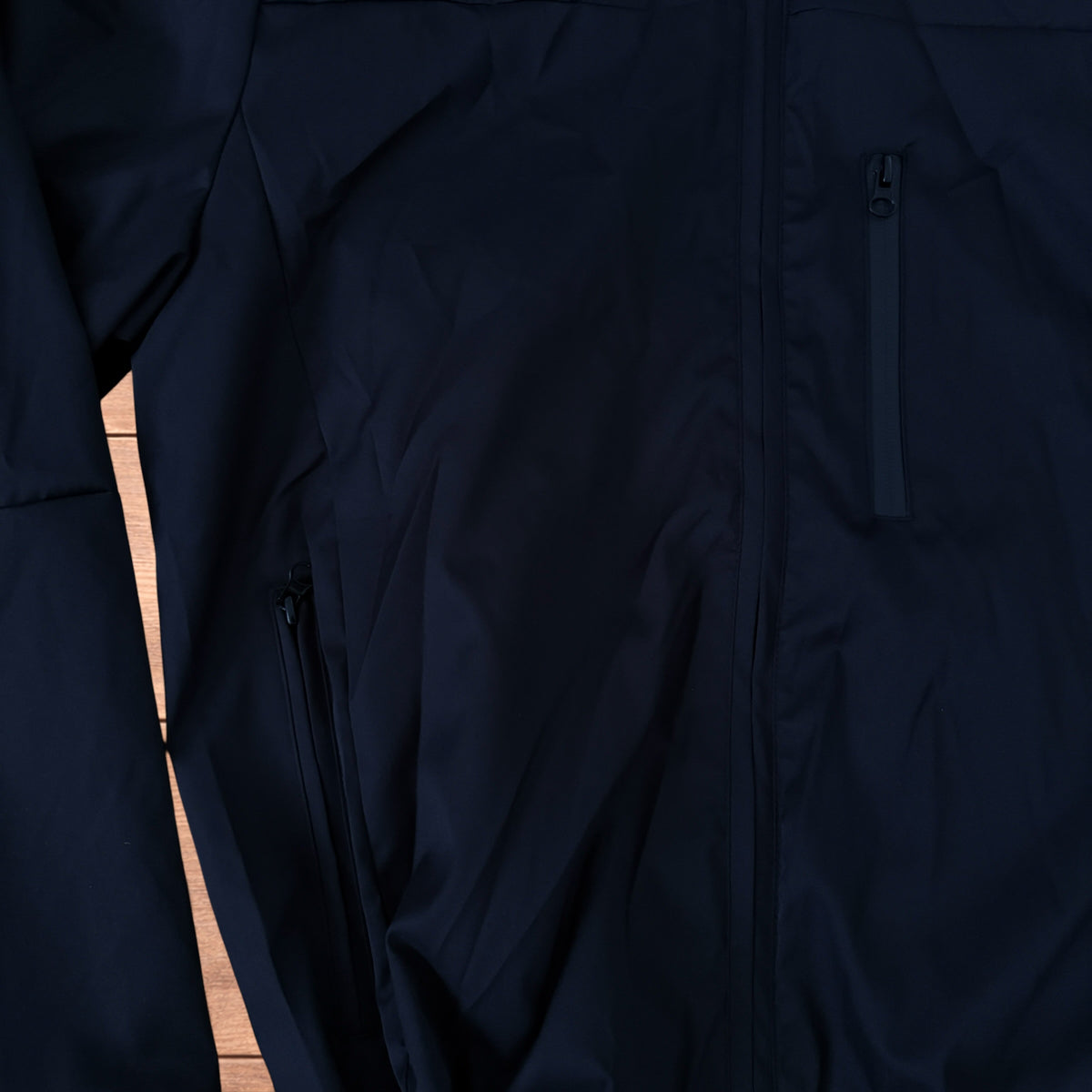 Black outdoor windbreaker jacket with hood, zip pockets and toggled hem laid flat on wooden surface.