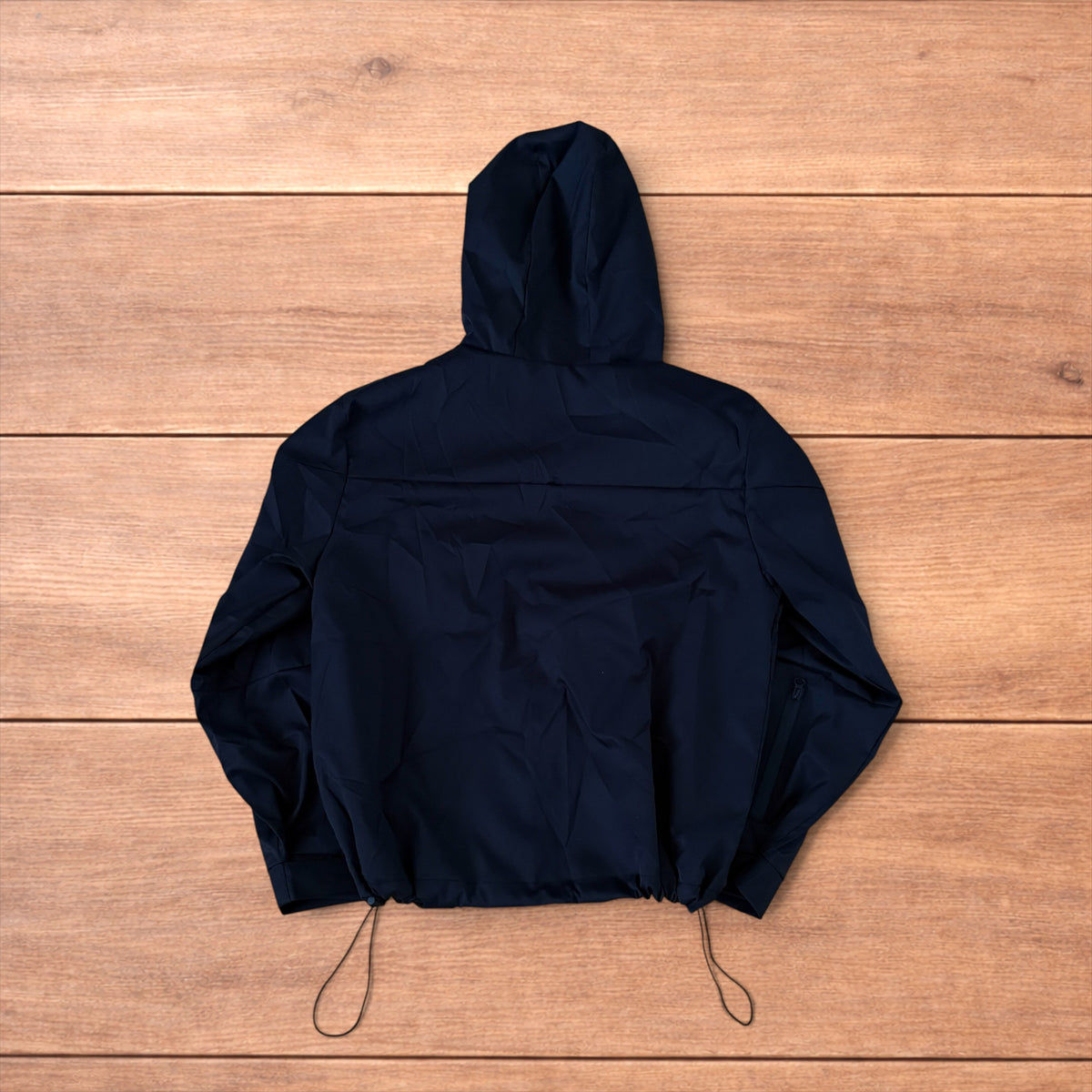 Black outdoor windbreaker jacket with hood, zip pockets and toggled hem laid flat on wooden surface.