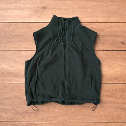 Dark green fleece utility gilet with zip pockets, drawstring neck, and toggled hem laid flat on wooden surface.