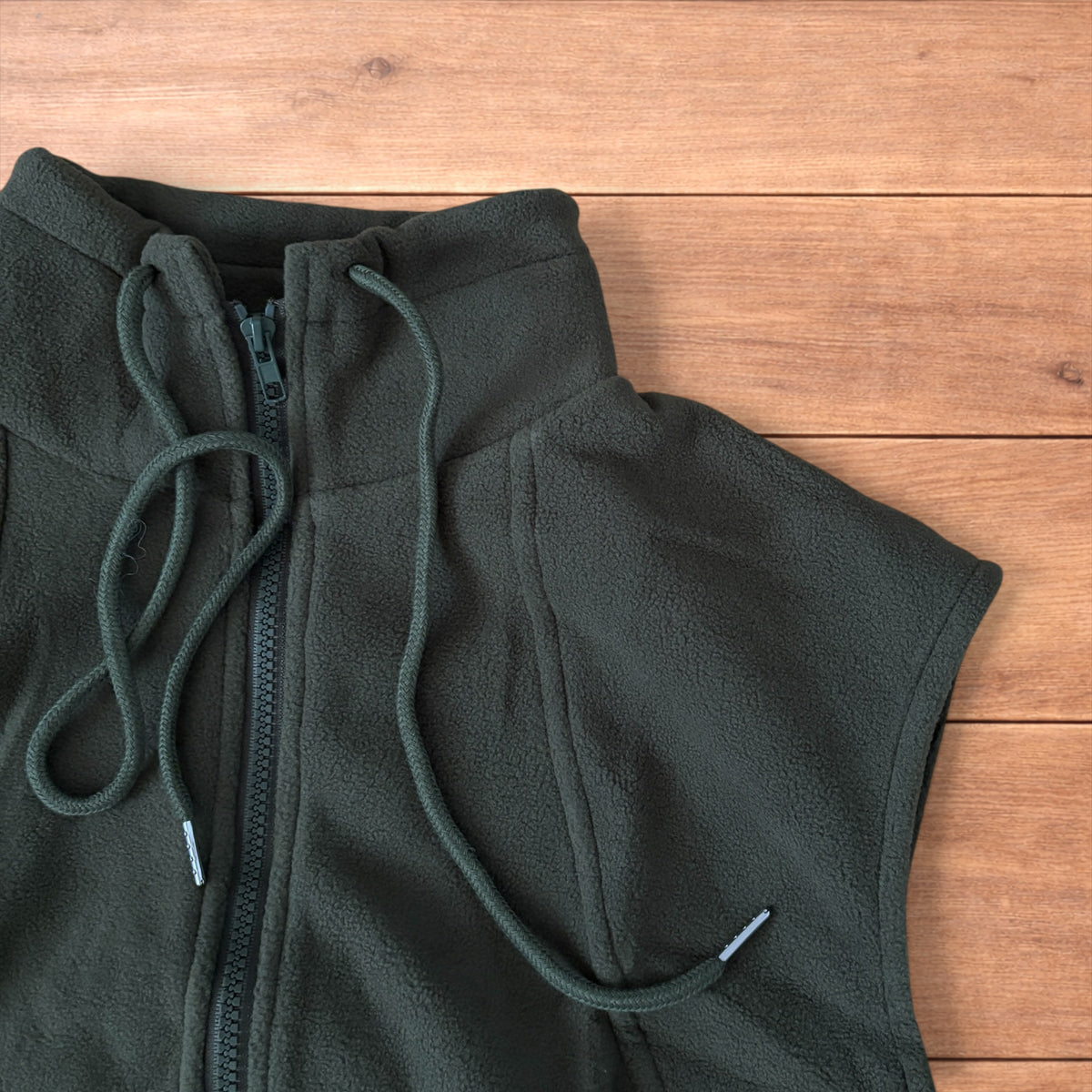 Dark green fleece utility gilet with zip pockets, drawstring neck, and toggled hem laid flat on wooden surface.