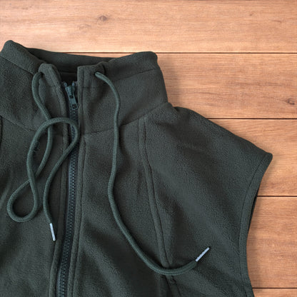 Dark green fleece utility gilet with zip pockets, drawstring neck, and toggled hem laid flat on wooden surface.
