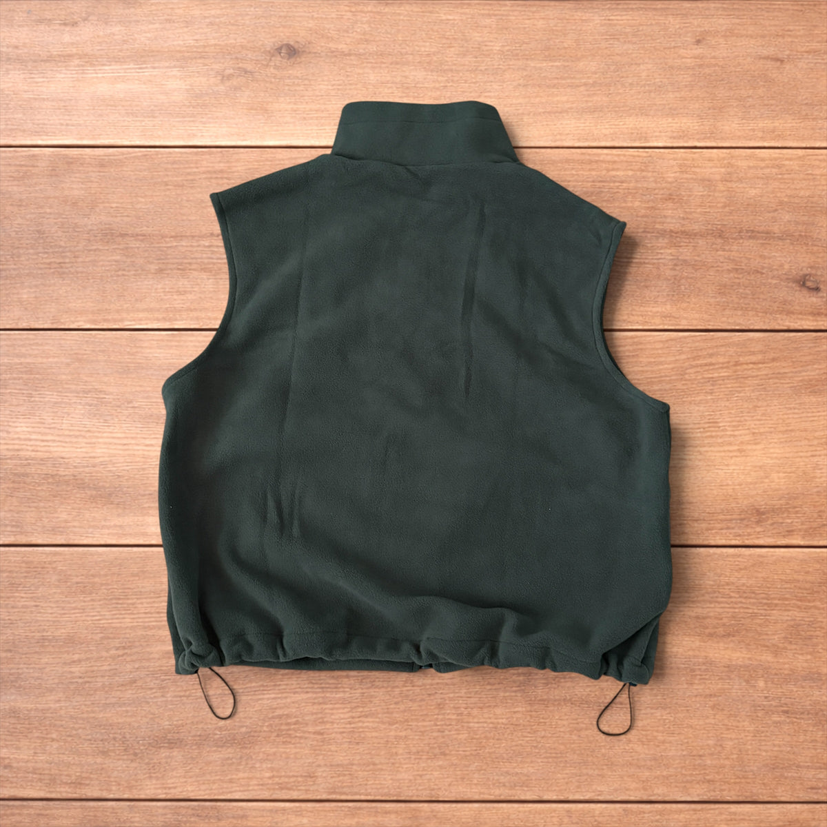 Dark green fleece utility gilet with zip pockets, drawstring neck, and toggled hem laid flat on wooden surface.