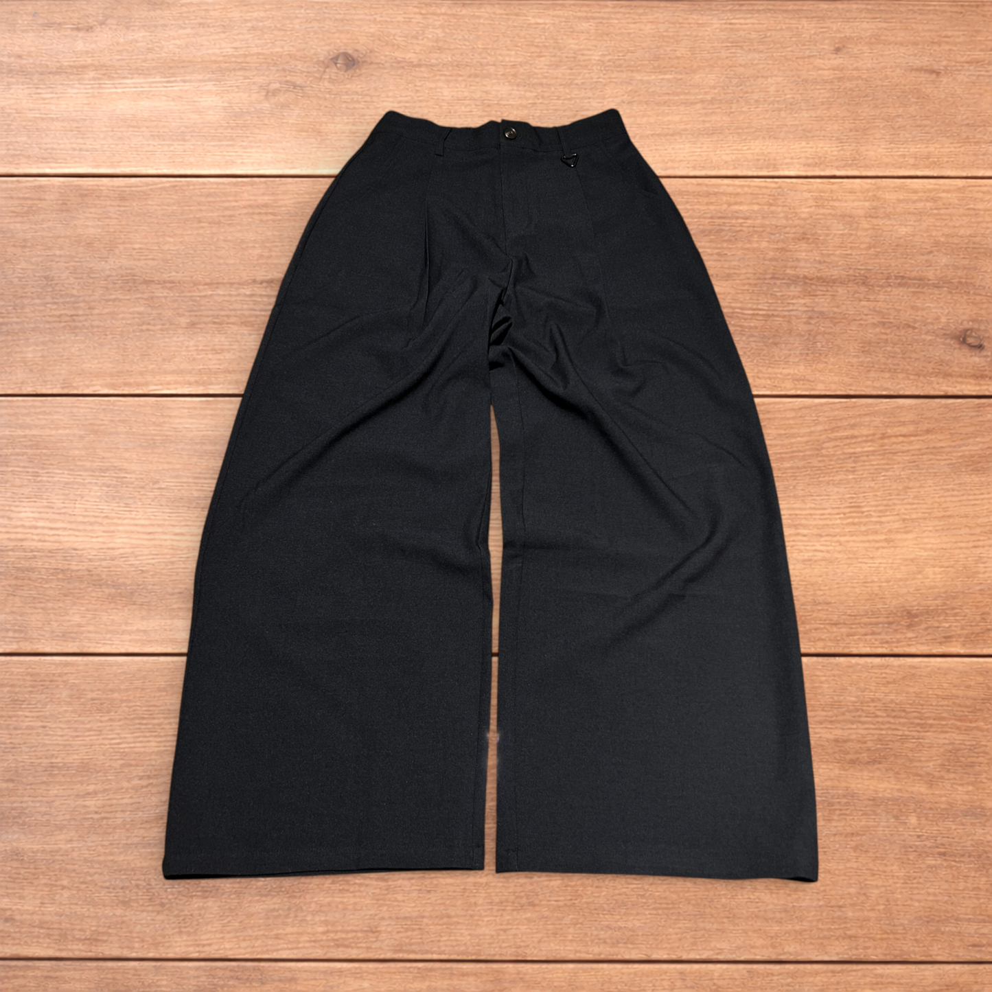 Black baggy wide leg tailored trousers with pleats and white stitch detail, laid flat on wood surface