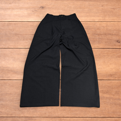 Black baggy wide leg tailored trousers with pleats and white stitch detail, laid flat on wood surface