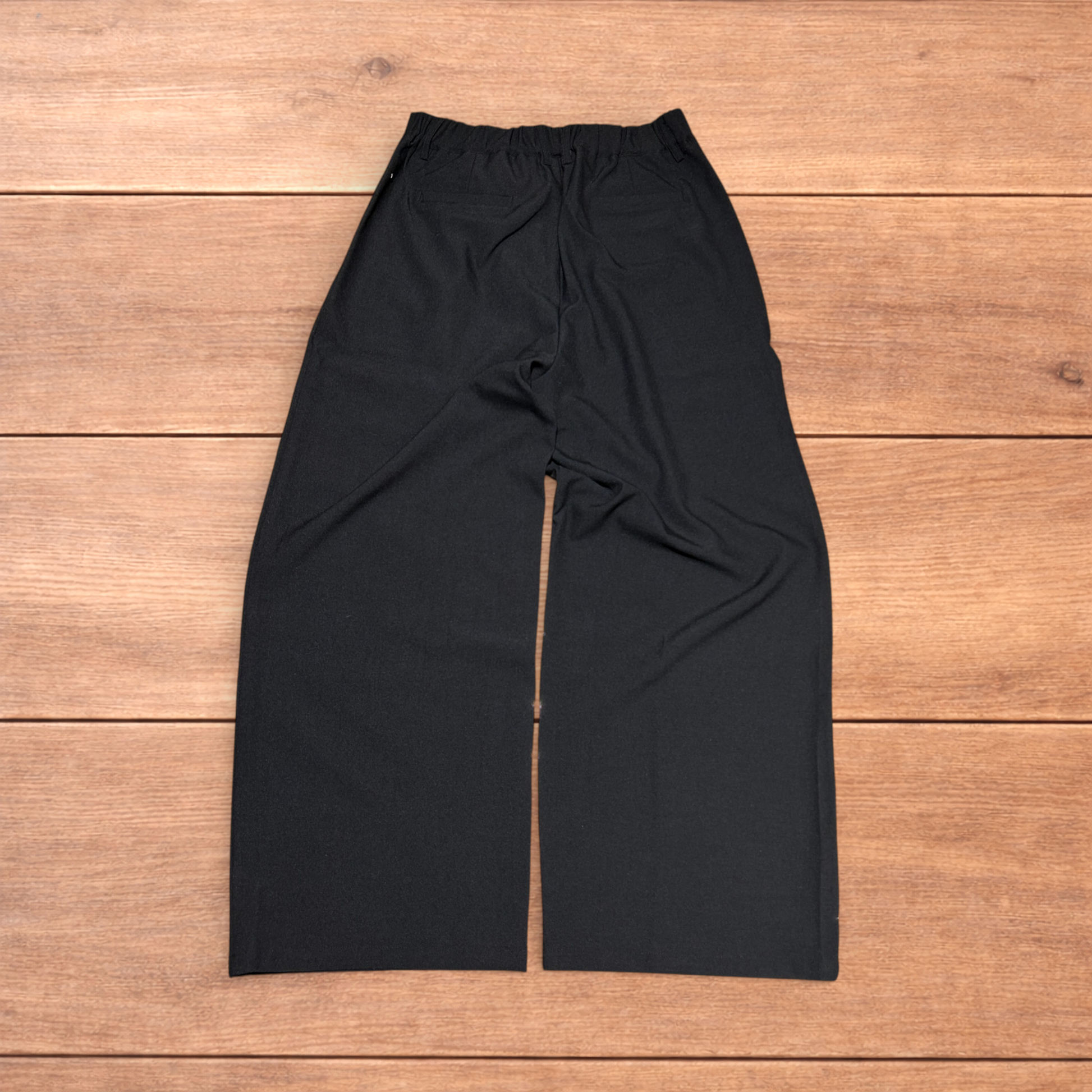 Black baggy wide leg tailored trousers with pleats and white stitch detail, laid flat on wood surface