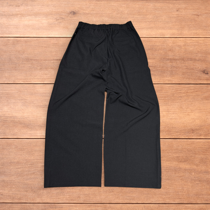 Black baggy wide leg tailored trousers with pleats and white stitch detail, laid flat on wood surface