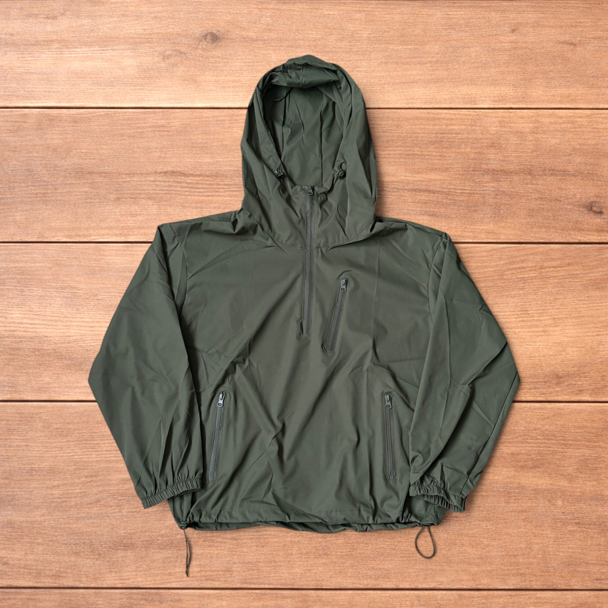 Khaki green lightweight shell anorak jacket with quarter zip and hood laid flat on wood surface.