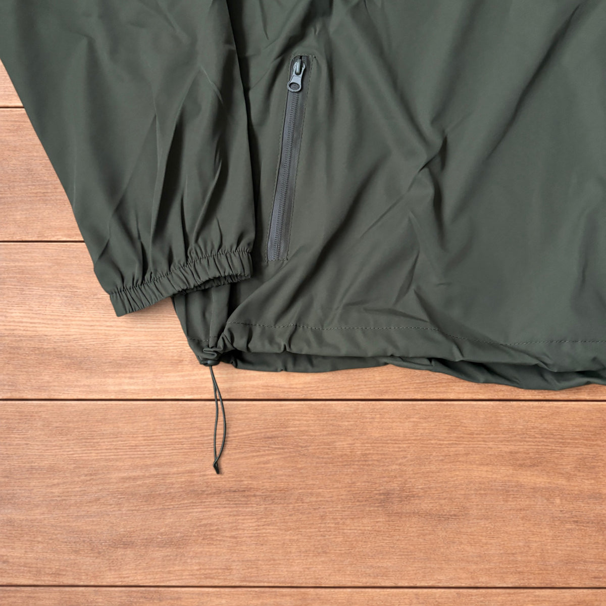 Khaki green lightweight shell anorak jacket with quarter zip and hood laid flat on wood surface.