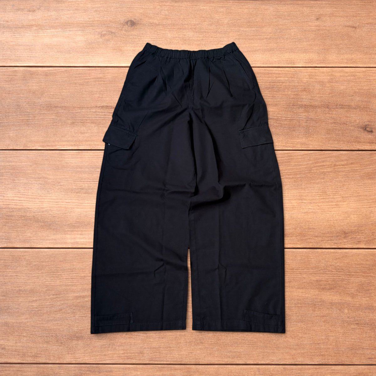 Black baggy wide leg cargo trousers laid flat on wooden surface showing elasticated waistband and cargo pockets.