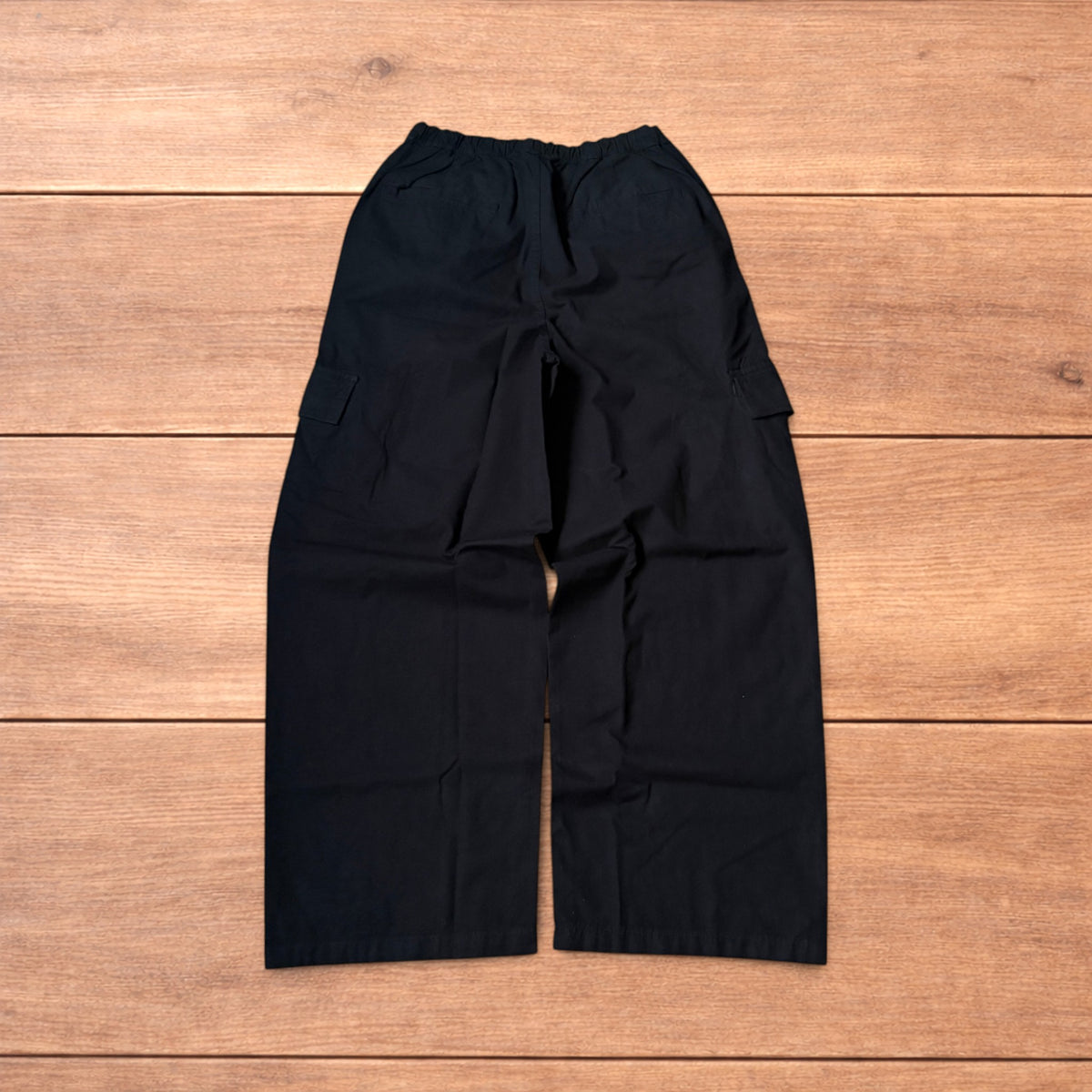 Black baggy wide leg cargo trousers laid flat on wooden surface showing elasticated waistband and cargo pockets.