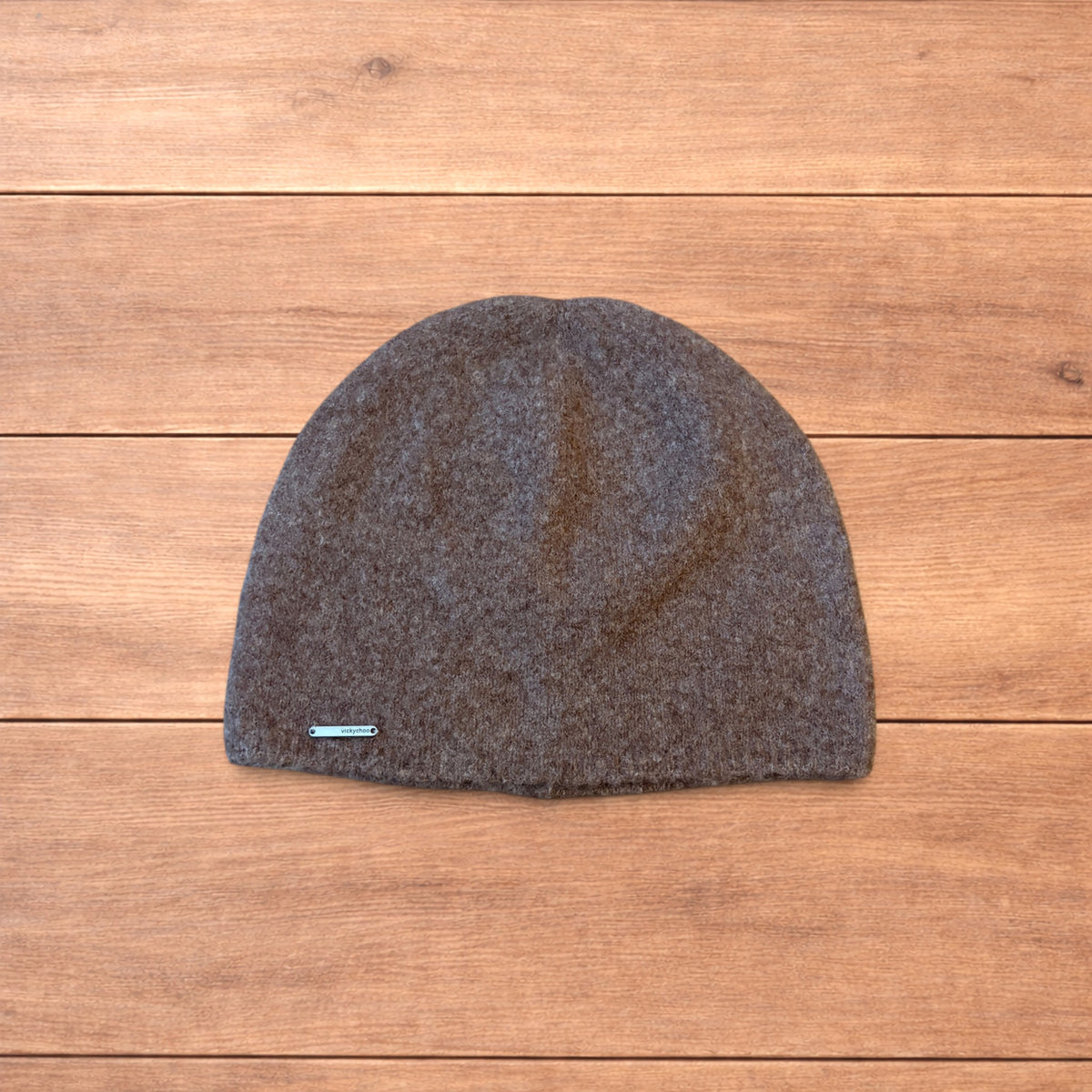 Pastel brown Korean style knitted beanie with sewn-on metal logo detail on wooden background.