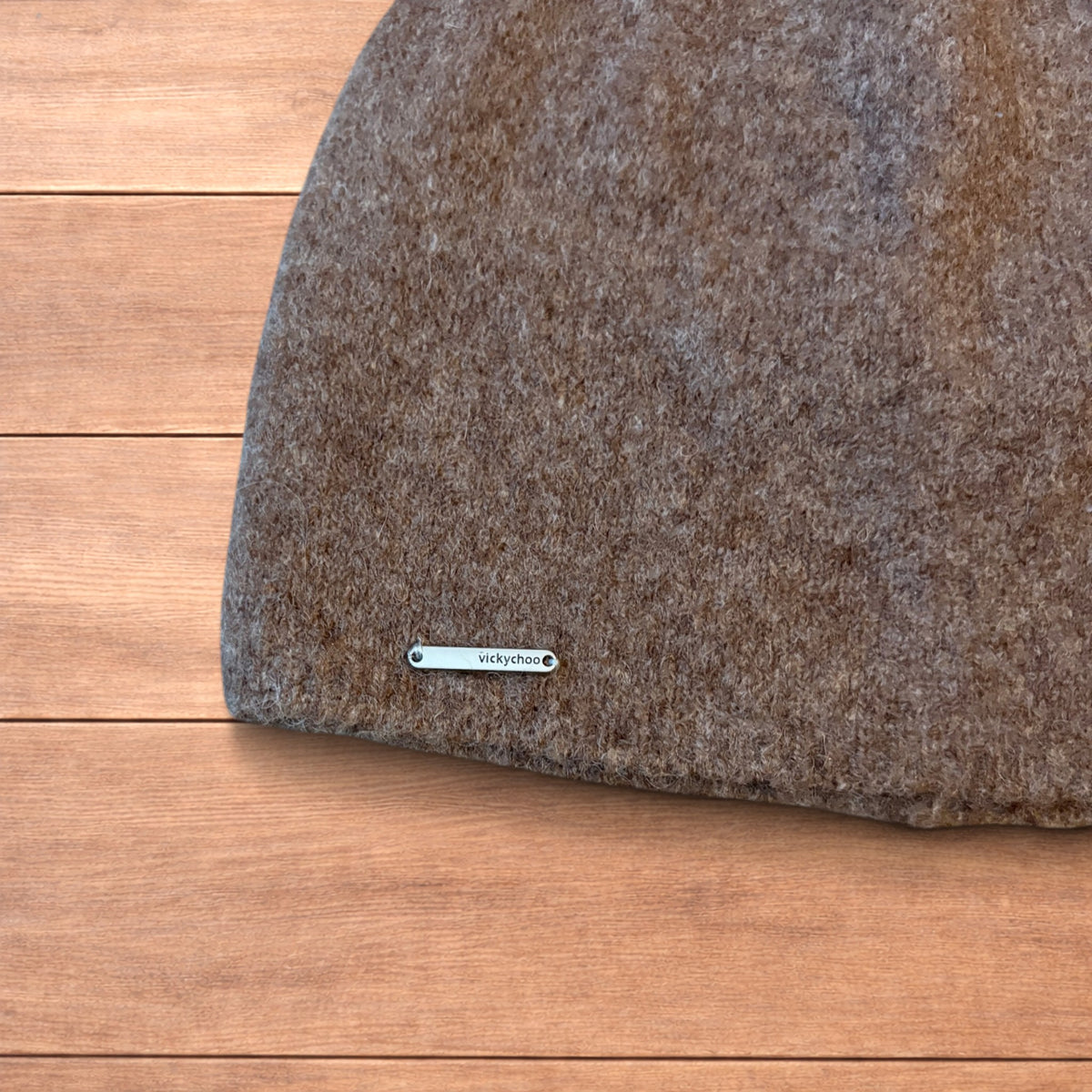 Pastel brown Korean style knitted beanie with sewn-on metal logo detail on wooden background.
