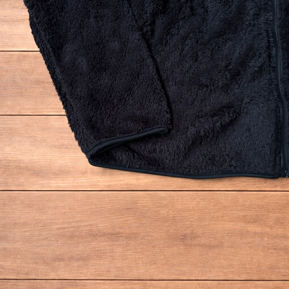 Black fleece fluffy zip up jacket laid flat on wooden floor showing full front, collar, sleeves, and pockets.