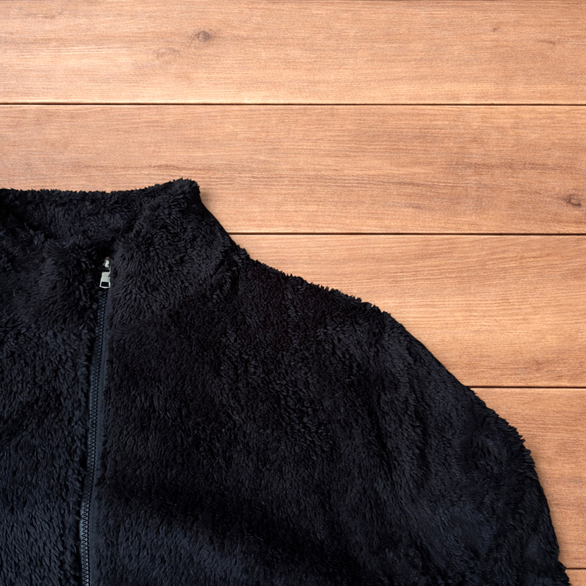 Black fleece fluffy zip up jacket laid flat on wooden floor showing full front, collar, sleeves, and pockets.