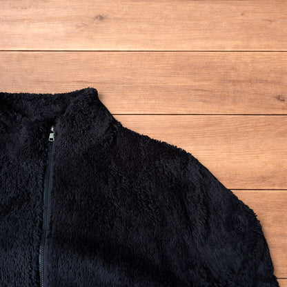 Black fleece fluffy zip up jacket laid flat on wooden floor showing full front, collar, sleeves, and pockets.