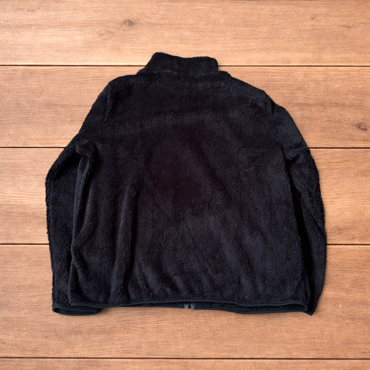 Black fleece fluffy zip up jacket laid flat on wooden floor showing full front, collar, sleeves, and pockets.