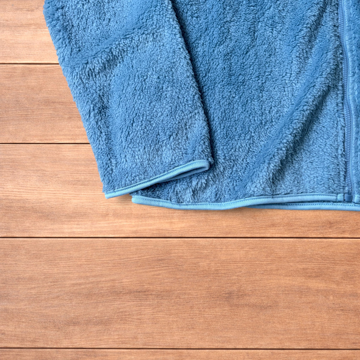 Light blue fleece fluffy zip up jacket laid flat on wooden surface.