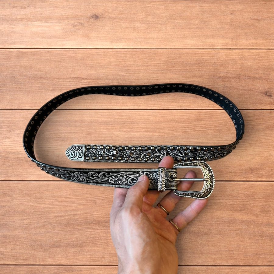 Black faux leather vintage style Western cowboy belt with silver embossed floral detailing and silver stud trim held in hand on wood background