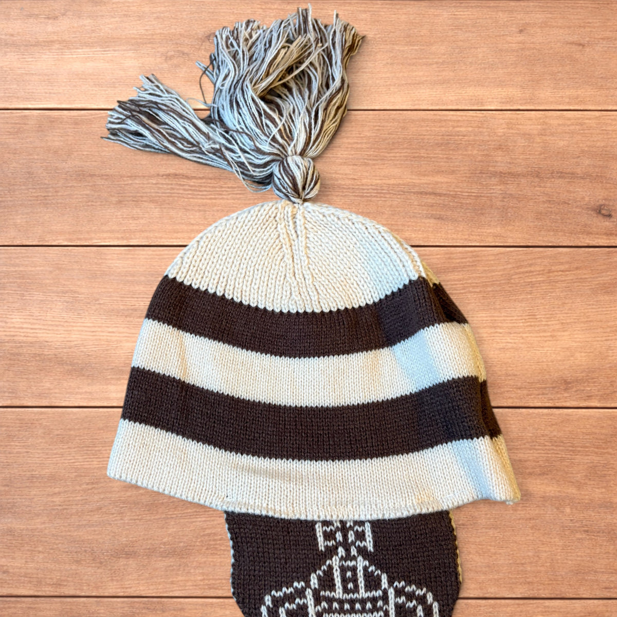 Vintage style striped dappy bobble hat with ear flaps and tassels in brown and cream laid flat on wooden surface.
