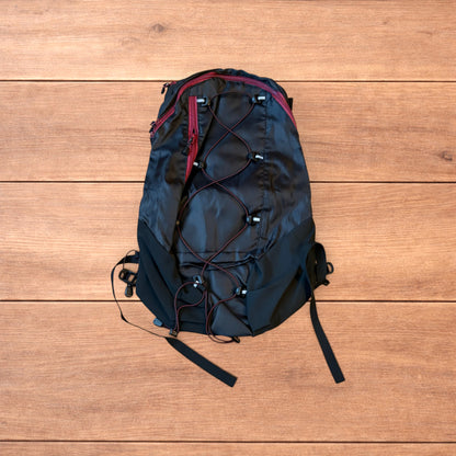 Black gorpcore outdoor hiking backpack with red zippers and elastic bungee cords laid flat on a wooden surface.