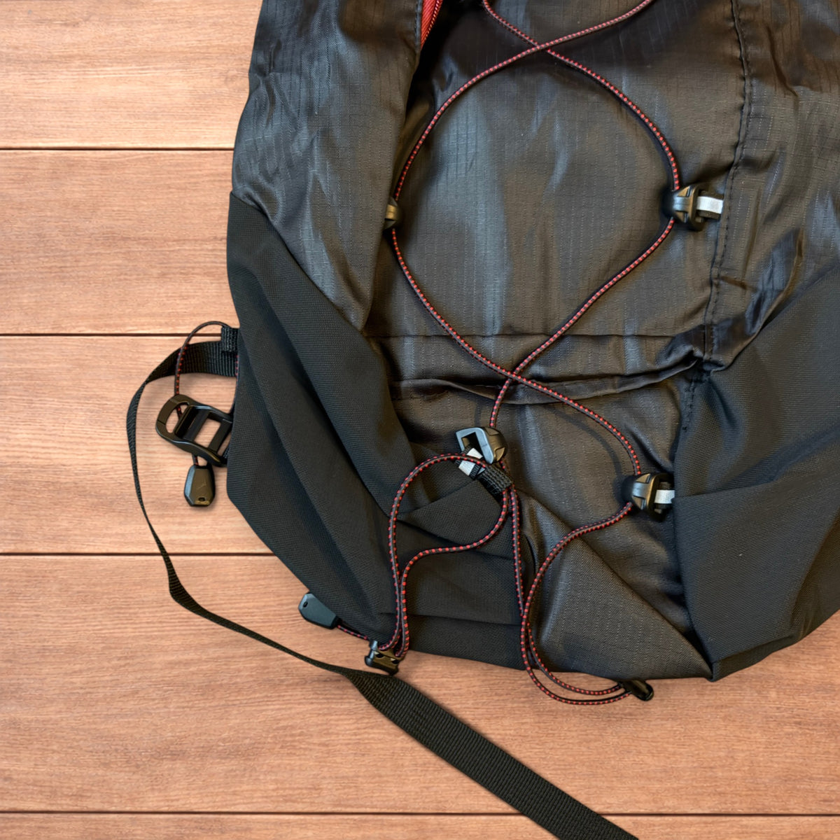 Black gorpcore outdoor hiking backpack with red zippers and elastic bungee cords laid flat on a wooden surface.
