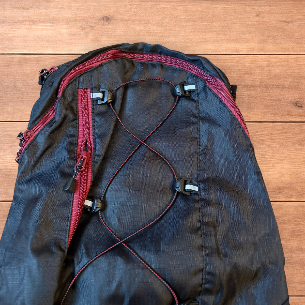 Black gorpcore outdoor hiking backpack with red zippers and elastic bungee cords laid flat on a wooden surface.