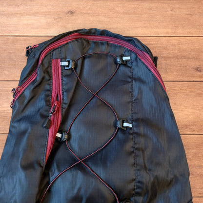 Black gorpcore outdoor hiking backpack with red zippers and elastic bungee cords laid flat on a wooden surface.