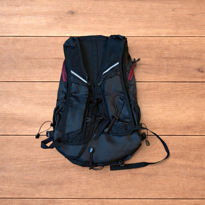 Black gorpcore outdoor hiking backpack with red zippers and elastic bungee cords laid flat on a wooden surface.