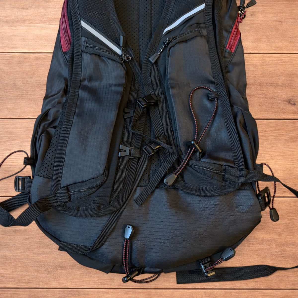 Black gorpcore outdoor hiking backpack with red zippers and elastic bungee cords laid flat on a wooden surface.