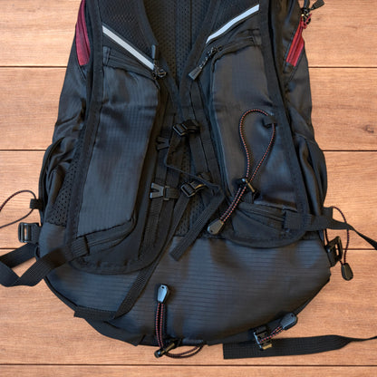 Black gorpcore outdoor hiking backpack with red zippers and elastic bungee cords laid flat on a wooden surface.