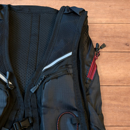 Black gorpcore outdoor hiking backpack with red zippers and elastic bungee cords laid flat on a wooden surface.