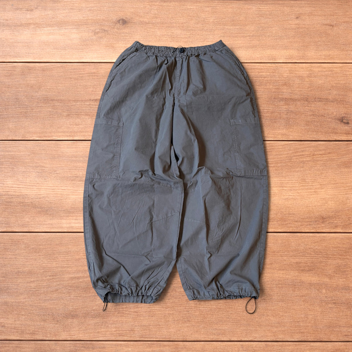 Grey baggy wide leg ripstop cargo trousers with cargo pockets, elasticated waistband and toggled hems laid flat on wooden floor.