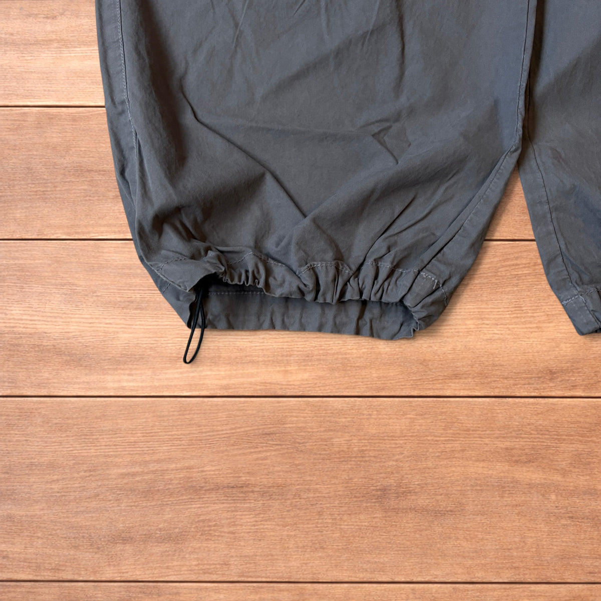 Grey baggy wide leg ripstop cargo trousers with cargo pockets, elasticated waistband and toggled hems laid flat on wooden floor.