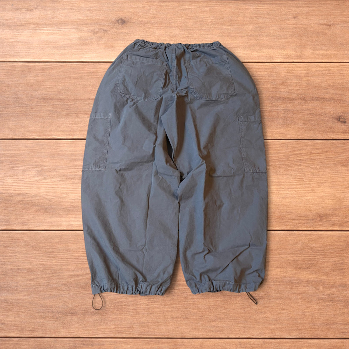 Grey baggy wide leg ripstop cargo trousers with cargo pockets, elasticated waistband and toggled hems laid flat on wooden floor.