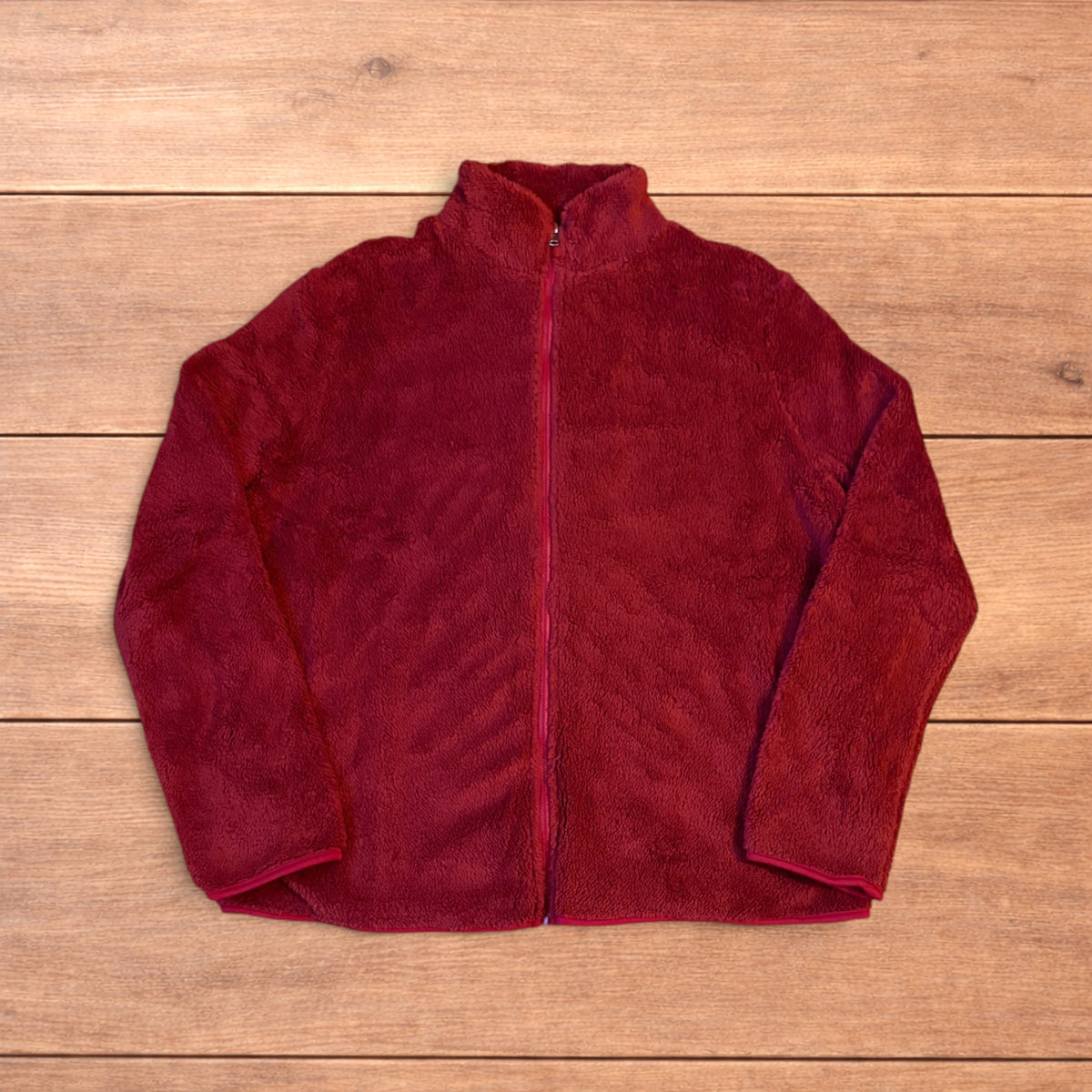 Deep dark red fluffy fleece zip up jacket laid flat on wooden surface.