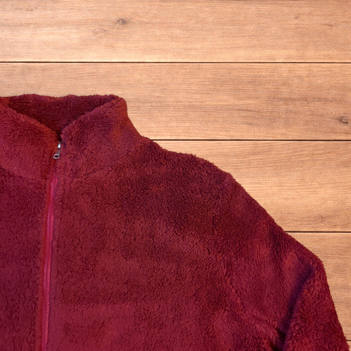 Deep dark red fluffy fleece zip up jacket laid flat on wooden surface.