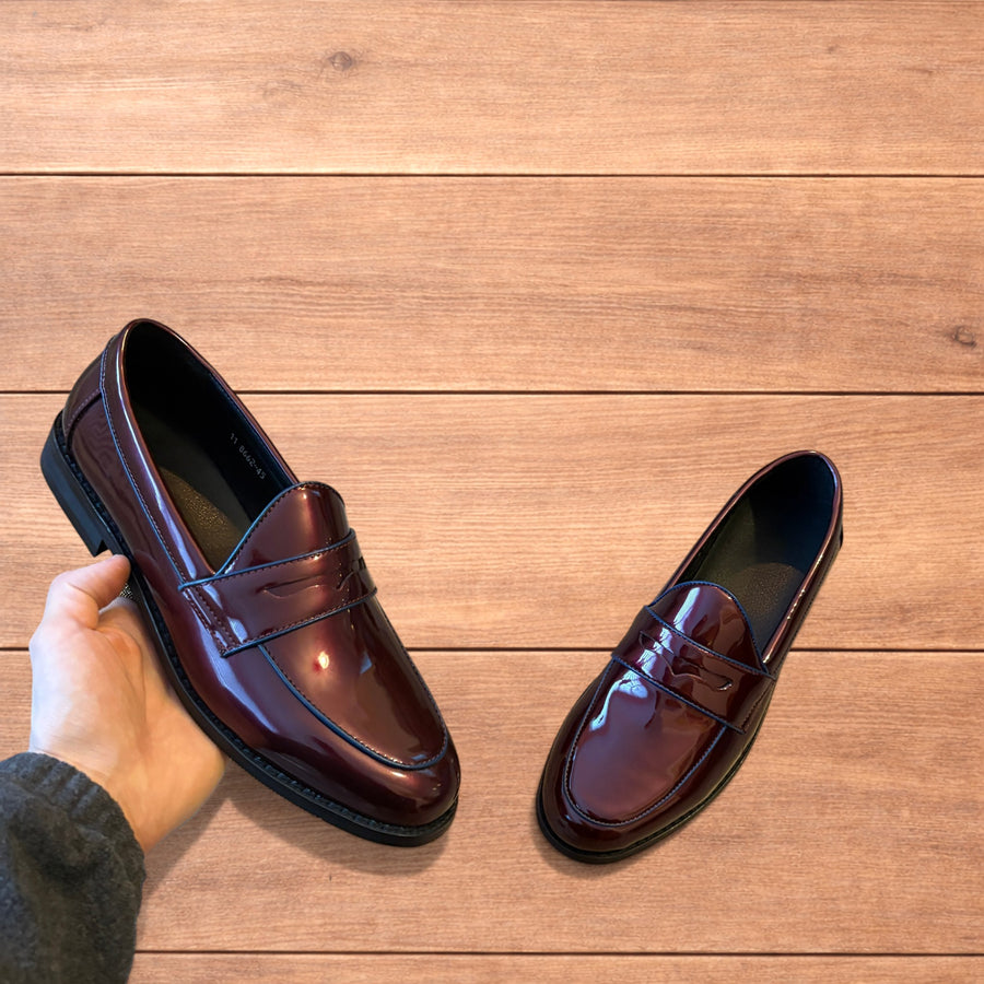 Pair of shiny oxblood red faux leather penny dress loafers held in hands on wooden floor