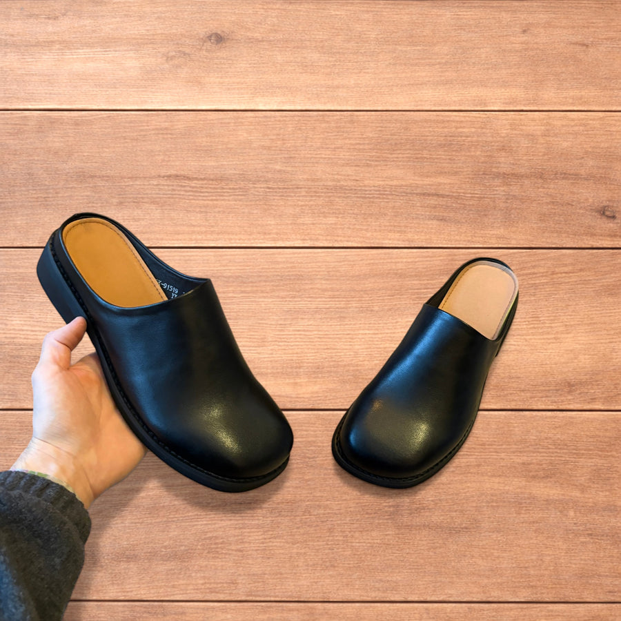 Black faux leather slip on clogs slippers with rounded toes and tan interior lining, shown from top and side views on wooden background