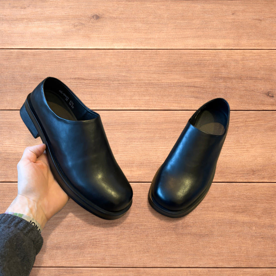 Pair of black faux leather slip-on loafers shoes on wooden floor, held in hands