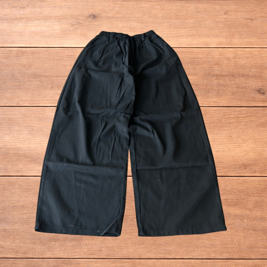 Black super baggy wide leg workwear trousers flat on wooden surface showing elasticated waistband and wide legs