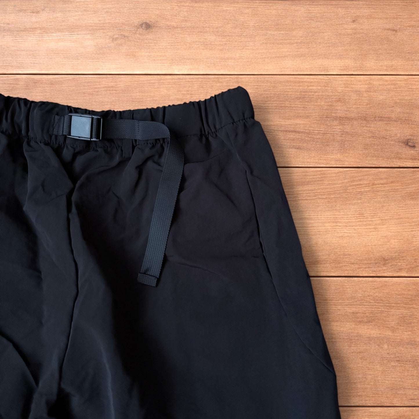 Baggy black gorpcore tech hiking pants with adjustable belted waist laid flat on wooden surface